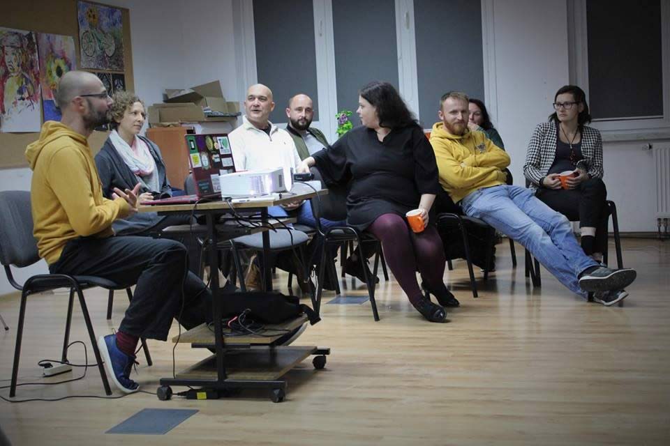 Na zdjęciu grupa osób siedzi na krzesłach w sali przypominającą szkolną. Pomiędzy nimi na stole stoi projektor multimedialny oraz laptop. Widać, że osoby rozmawiają ze sobą. 